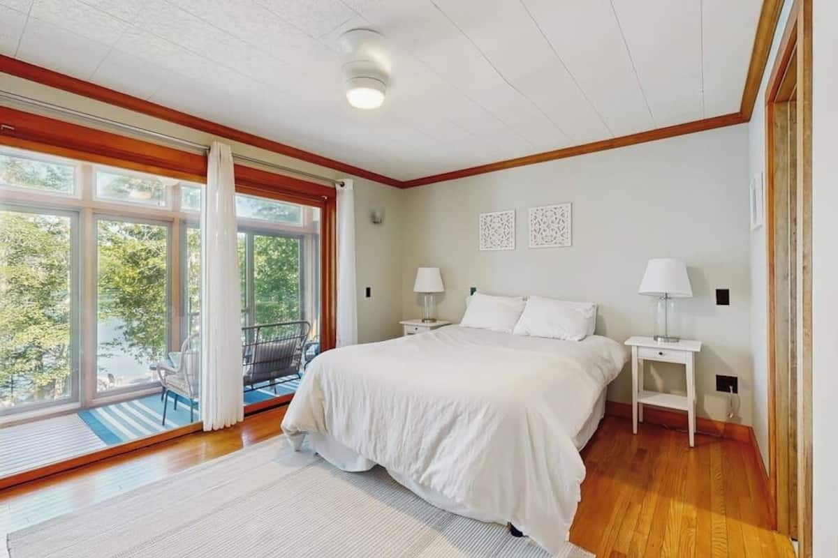 A bedroom with a large window, a bed, two bedside tables with lamps, and a view of greenery outside.
