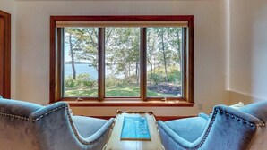 Ein Zimmer mit großem Fenster und Blick auf Bäume sowie ein Gewässer. Zwei Polstersessel und ein kleiner Tisch mit einem Buch sind vorhanden.