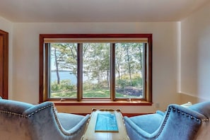 A room with a large window offering a view of trees and a body of water. There are two upholstered chairs and a small table with a book. - Delightful Oceanfront Home With Amazing Views, Wrap-around Deck & two Fireplaces (Wiscasset)