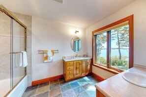 Ein Badezimmer mit Duschkabine aus Glas, einem Holzbadezimmermöbel mit Spiegel und einem großen Fenster mit Blick ins Freie.