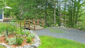 En træbro over en grussti, omgivet af en have med forskellige planter og en træterrasse med pergola.
