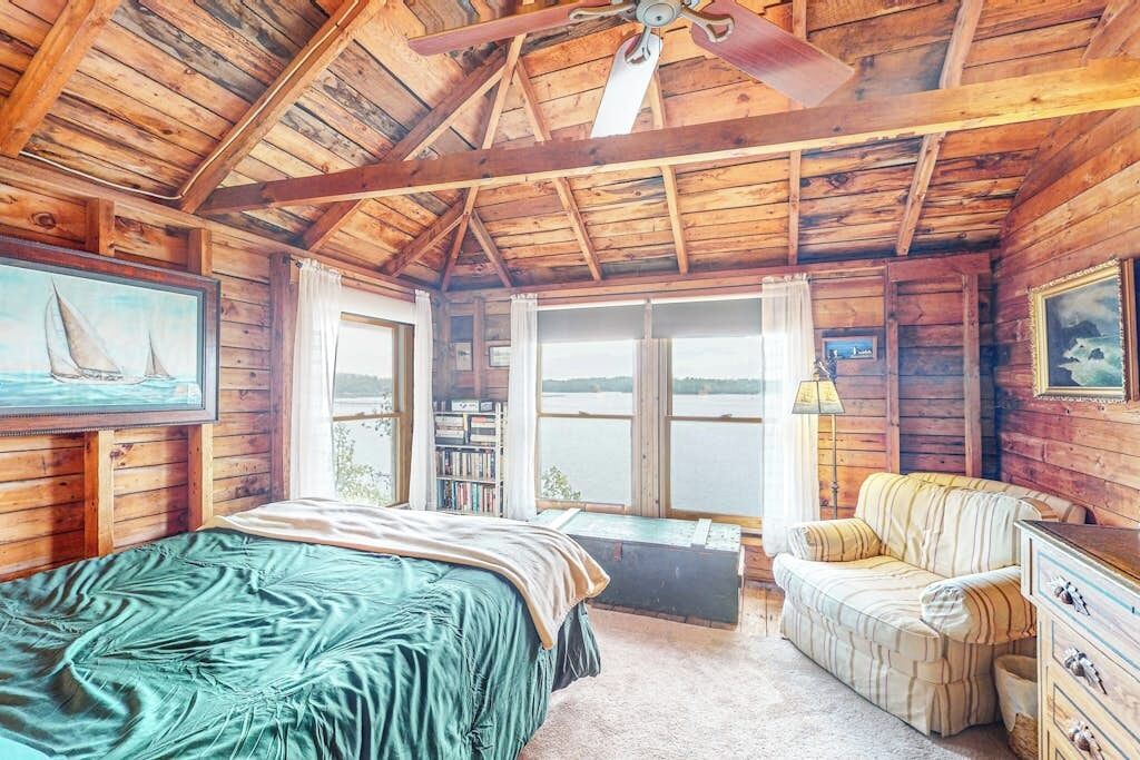 A wooden cabin bedroom with a bed, a sofa, a ceiling fan, and a view of a lake.