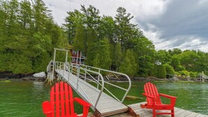 A wooden dock with two red Adirondack chairs, a metal bridge, and a house in the background. - Cozy, Waterfront Cabin-style Gem With Private Dock and Lovely Views (Bath)