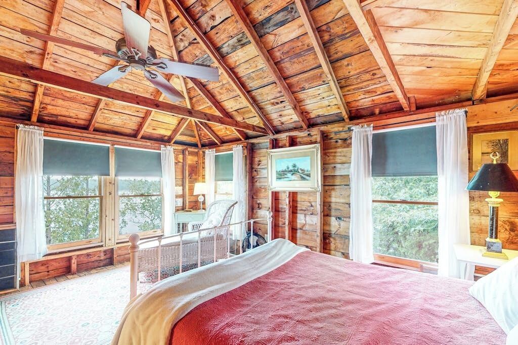 Un dormitorio de cabaña de madera con una cama, ventilador de techo y ventanas amplias.