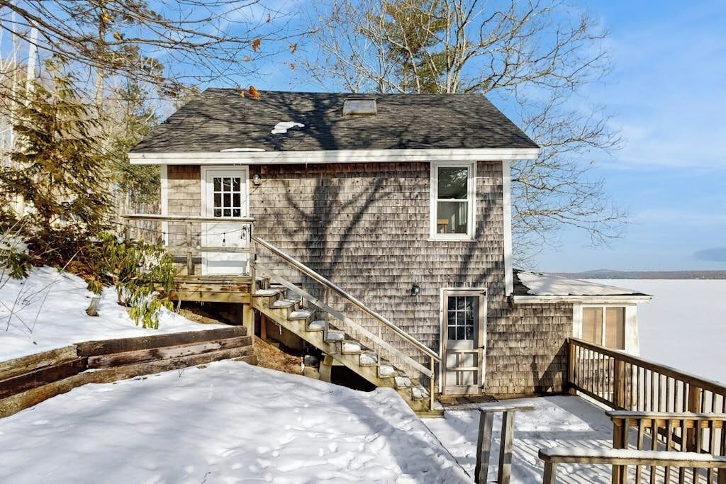 A wooden house with a staircase leading to a deck, surrounded by snow and trees.