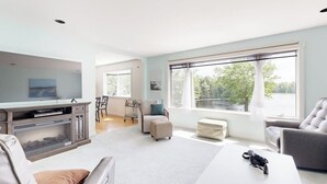 A spacious living room with a television, a fireplace, a dining area, and large windows with a view. - Charming Lakefront House With Outdoor Firepit, Washer/dryer and Wifi (Gouldsboro)