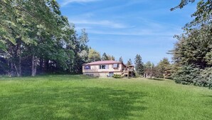 A two-story house with a red roof, surrounded by a large green lawn and dense trees. - Charming Lakefront House With Outdoor Firepit, Washer/dryer and Wifi (Gouldsboro)