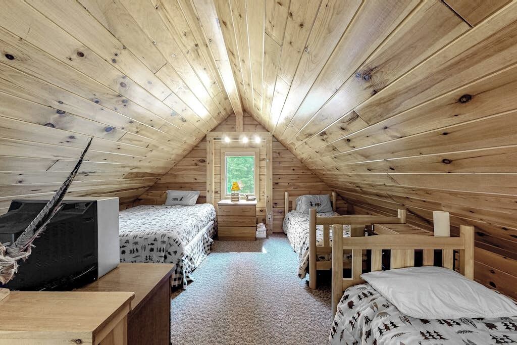A cozy attic bedroom with wooden walls and sloped ceilings, featuring two beds, a desk, and a window with a view.