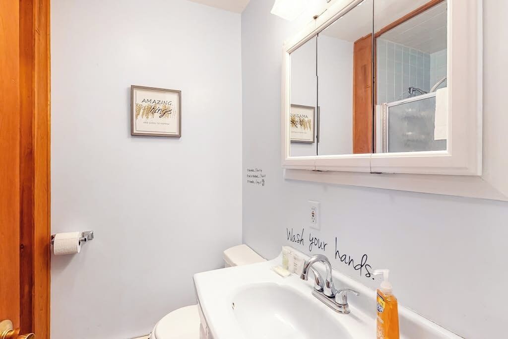 Un baño con un lavamanos blanco, un espejo, una cita enmarcada en la pared y un cartel que dice 'Lávate las manos'.