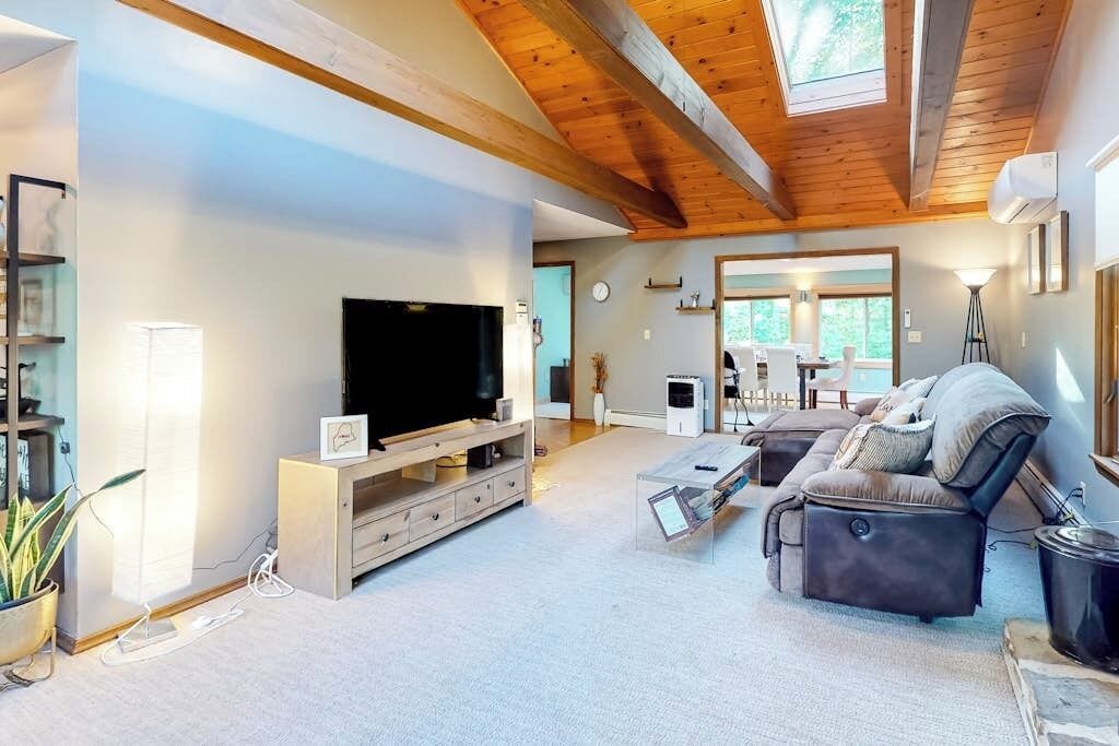 A modern living room with a large flat-screen TV, a sofa, a coffee table, and a wooden ceiling.