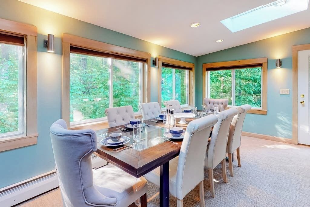 A dining area with a long table, six chairs, and large windows offering a view of greenery.