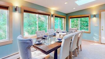 A dining area with a long table, six chairs, and large windows offering a view of greenery.