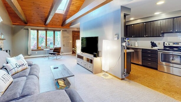 A modern living room with a sofa, a coffee table, and a TV. There is a kitchen area with cabinets and appliances. - Tree-lined Home With Screened Porch & Perfect Midcoast Location (Damariscotta)