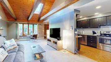 A modern living room with a sofa, a coffee table, and a TV. There is a kitchen area with cabinets and appliances.