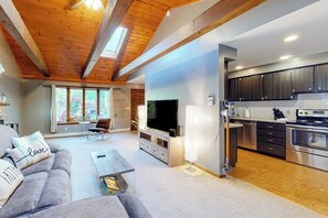 Una sala de estar moderna con un sofá, una mesa de centro y un televisor. Se observa un área de cocina con armarios y electrodomésticos.