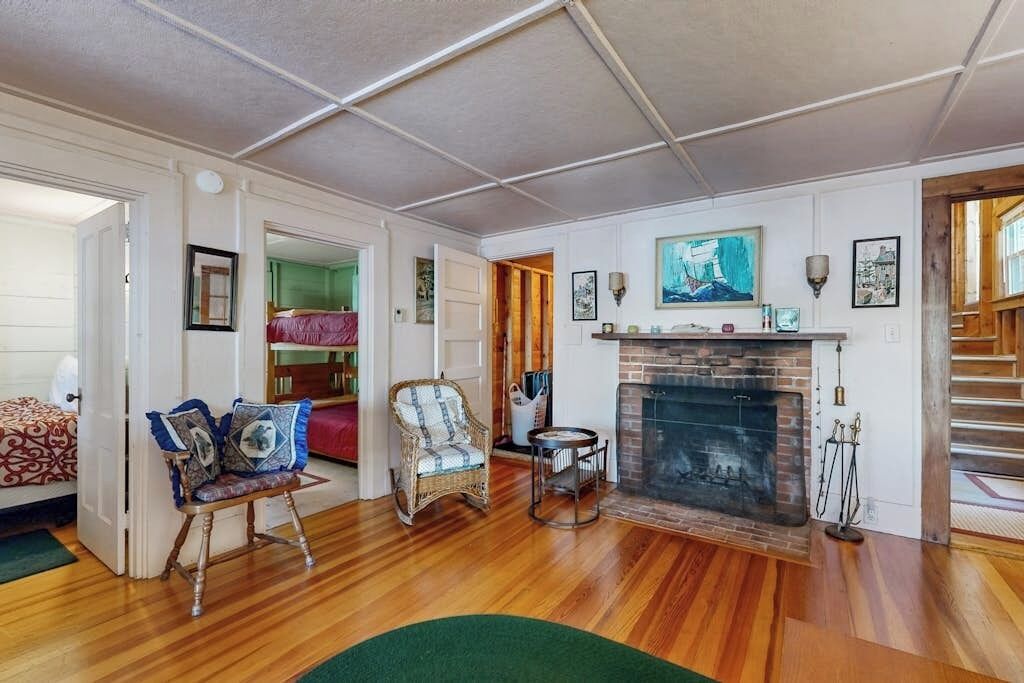 A living room with a fireplace, wooden flooring, a green rug, wicker chairs, and a bed with patterned bedding.