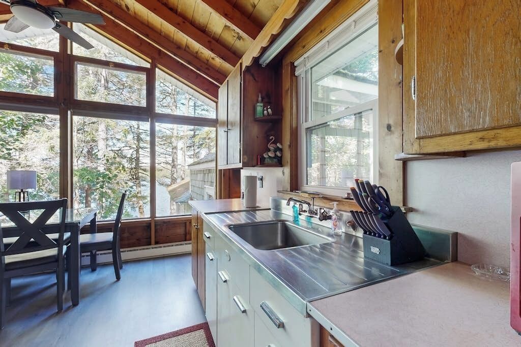 A kitchen with a stainless steel sink, wooden cabinets, and a window overlooking a forest.
