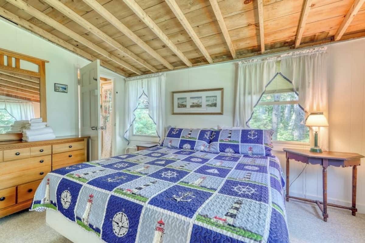 Un dormitorio con techo de madera, una cama con colcha de temática náutica, un aparador, una mesita de noche y una ventana con cortinas.