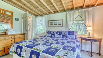 A bedroom with a wooden ceiling, a bed with a nautical-themed quilt, a dresser, a nightstand, and a window with curtains.