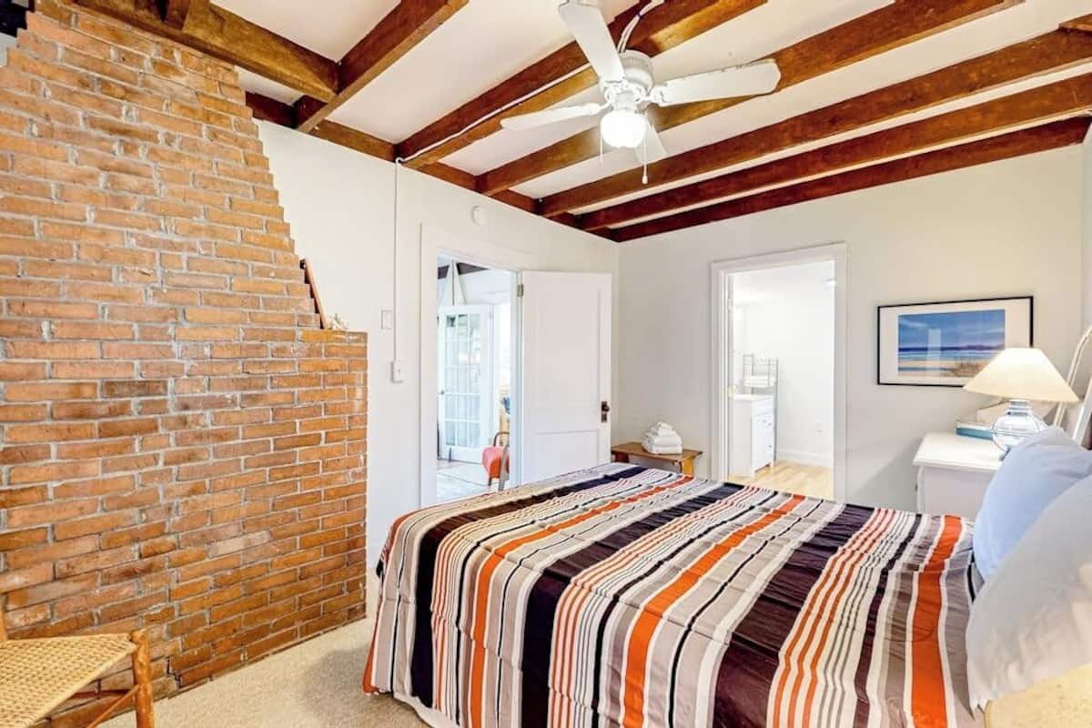 Un dormitorio con pared de ladrillo, vigas de madera, colcha a rayas, ventilador de techo y puerta que da a otra habitación.