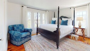 Quarto com cama de dossel, uma poltrona azul estampada, uma pequena mesa de cabeceira e uma janela com cortinas.