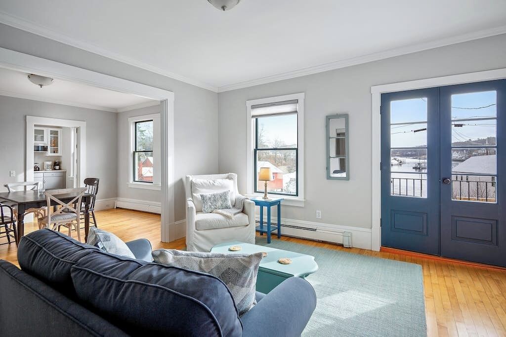 A modern living room with a sofa, armchair, dining table, and chairs. There are windows with views of a cityscape and a door with a balcony.