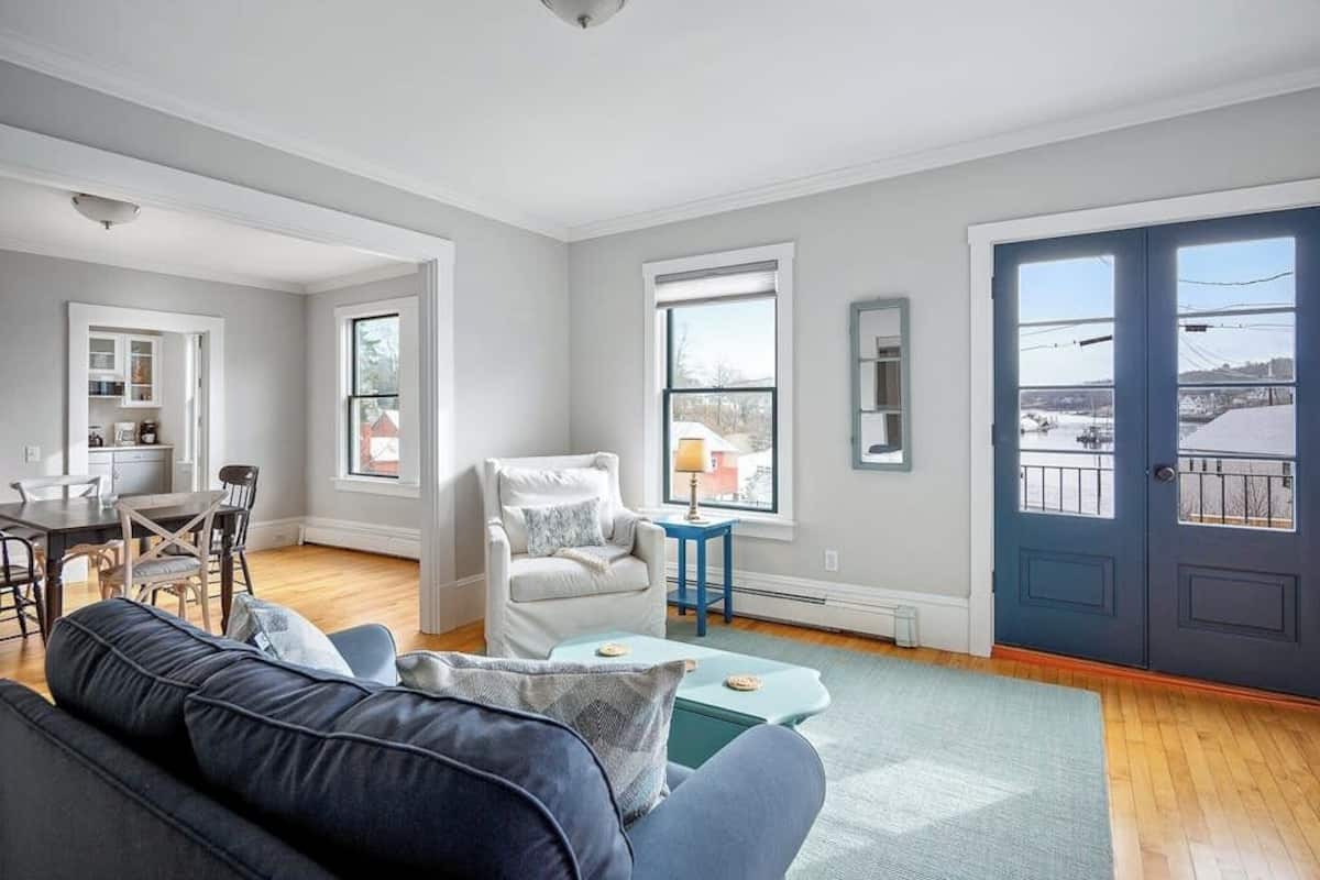 Una sala de estar moderna con sofá, sillón, mesa de comedor y sillas. Se ven ventanas con vistas a un paisaje urbano y una puerta con balcón.