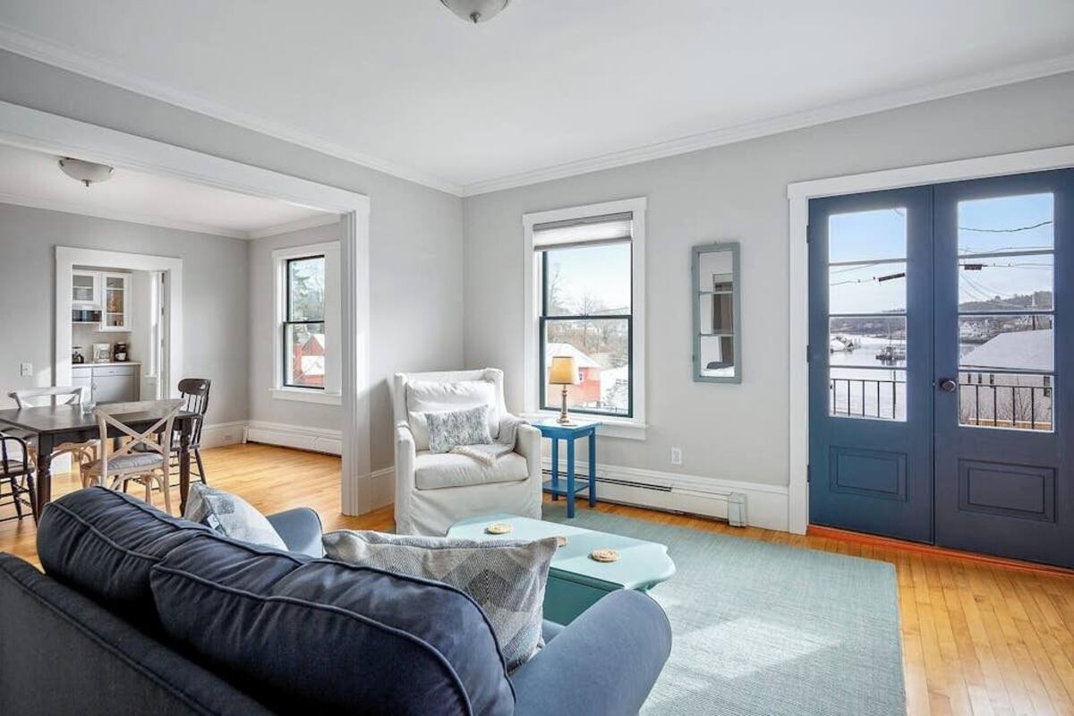 A modern living room with a sofa, armchair, dining table, and chairs. There are windows with views of a cityscape and a door with a balcony.