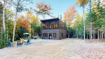 A modern log cabin with a dirt driveway, surrounded by trees and a fire pit area.