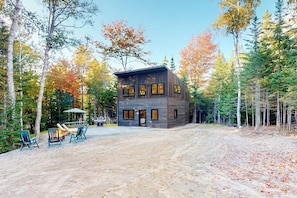 A modern log cabin with a dirt driveway, surrounded by trees and a fire pit area. - Partridge Cove (Ellsworth)