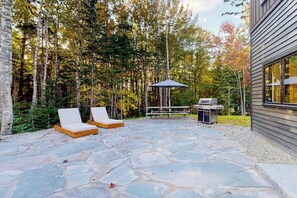 A stone patio with lounge chairs, a grill, and a gazebo in a forested area. - Partridge Cove (Ellsworth)