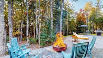 A forest setting with a fire pit, outdoor seating, and a picnic table.