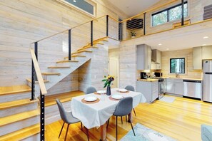 A modern kitchen with wooden floors, a dining area with a table set for four, and a staircase with a glass railing. - Partridge Cove (Ellsworth)