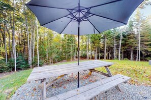 A wooden picnic table with an umbrella in a forested area. - Partridge Cove (Ellsworth)