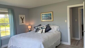 A bedroom with a bed, a window with curtains, a wall-mounted picture, and a door. - Abenaki Retreat on Matinicus Island (Matinicus)