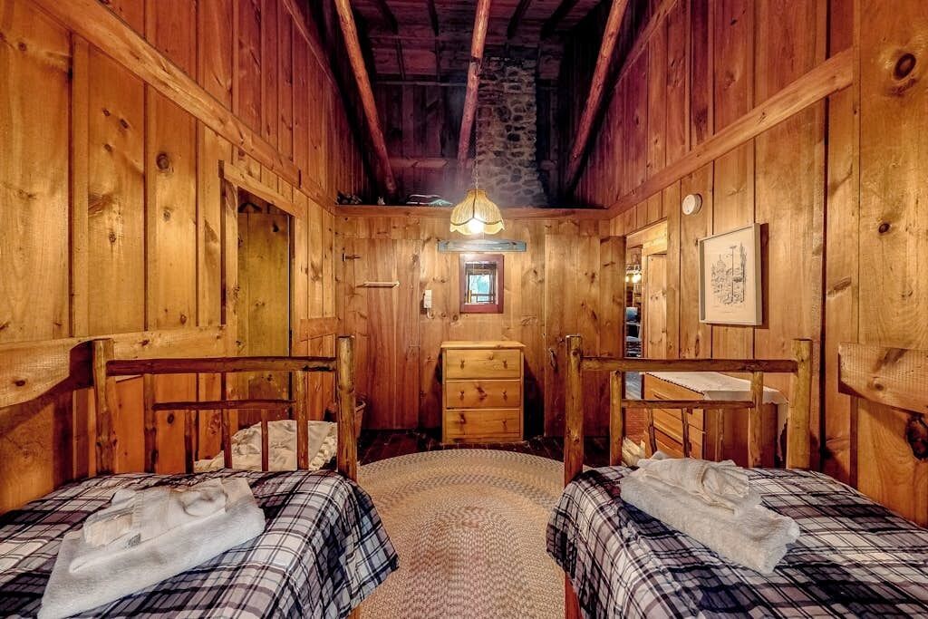Une pièce de style cabane en bois, avec deux lits, une commode en bois, une petite fenêtre et un luminaire suspendu.