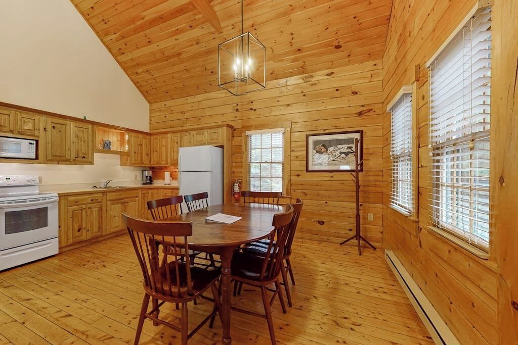 A wooden cabin interior with a kitchen, dining table, chairs, and a standing lamp.
