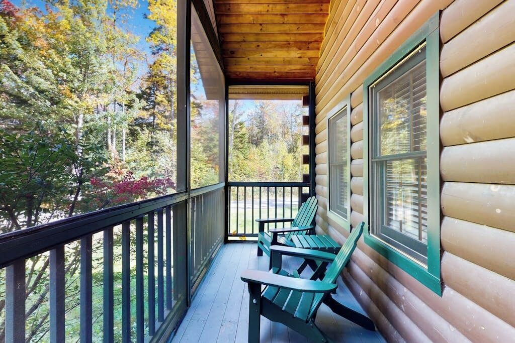 A wooden cabin with a porch, two Adirondack chairs, and a view of autumn trees.