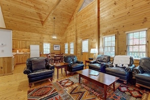 A log cabin interior with a dining area, leather sofas, a coffee table, and a kitchen in the background. - Point Lookout East (Lincolnville)