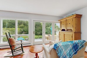 Ein Zimmer mit Schaukelstuhl, einer Couch mit kariertem Überwurf, einem Holzschrank und großen Fenstern mit Blick ins Grüne.