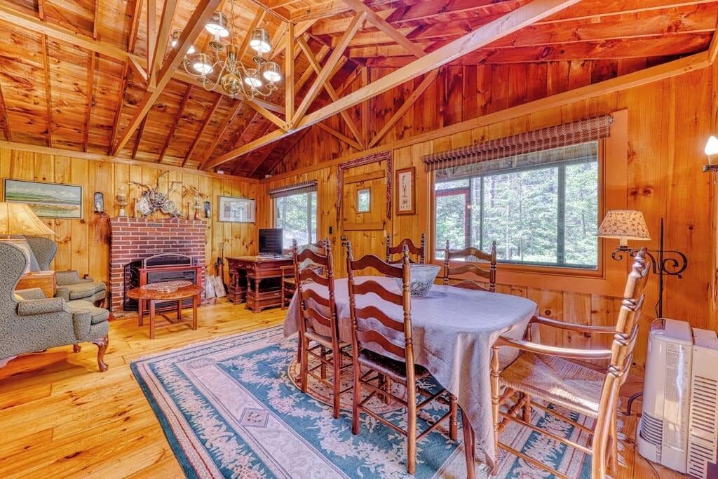 A wooden cabin interior with a dining area, fireplace, and large windows.