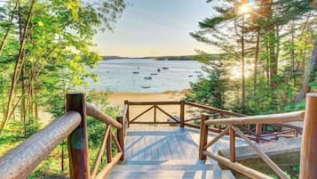 A wooden deck leading to a beach with boats, surrounded by trees and a clear sky.