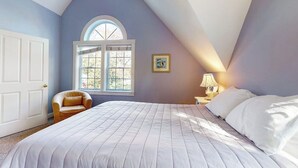 A bedroom with a large bed, a window with a view, a chair, and a nightstand with a lamp. - Harbor House (Camden)