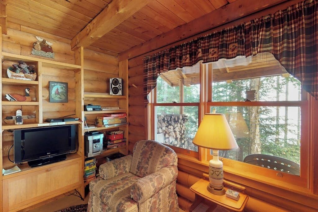 A wooden cabin interior with a television, shelves with various items, a plaid window valance, and a lamp.