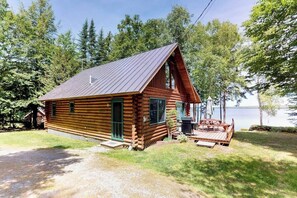 Una cabaña de troncos con porche y terraza, rodeada de árboles y un lago.