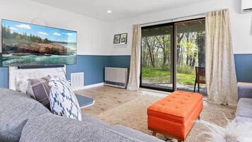 Uma sala de estar moderna com uma televisão de tela plana grande, um sofá, um banquinho e uma porta de vidro deslizante que dá para um jardim.