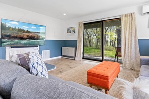 Uma sala de estar moderna com uma televisão de tela plana grande, um sofá, um banquinho e uma porta de vidro deslizante que dá para um jardim.
