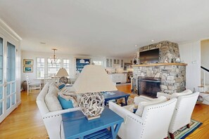 Ein geräumiges Wohnzimmer mit Kamin aus Stein, weißen Ledersofas, einem blauen Couchtisch und einem an den Kamin montierten Fernseher.