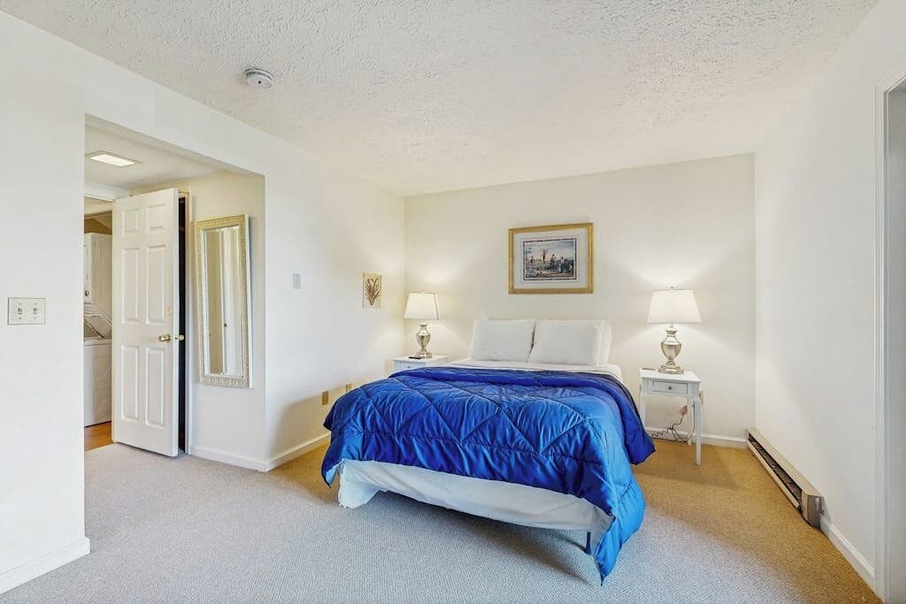 A bedroom with a bed, two bedside lamps, a picture on the wall, and a door leading to another room.