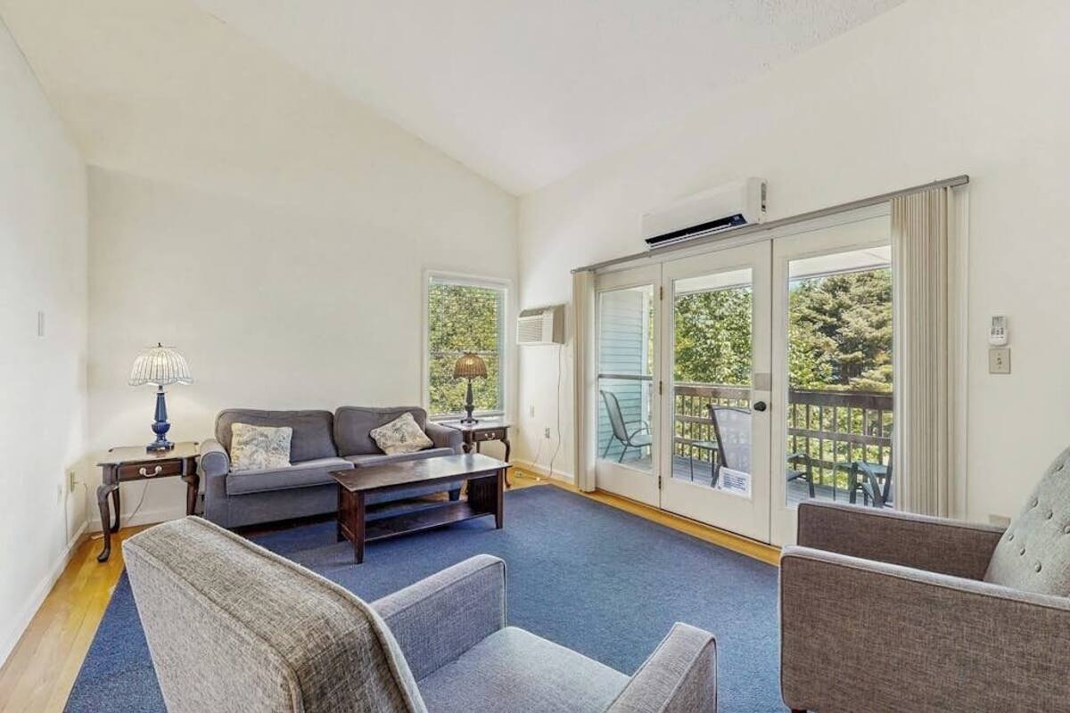 A living room with a sofa, chairs, a coffee table, and a lamp. There is a sliding glass door leading to an outdoor area with a view of trees.
